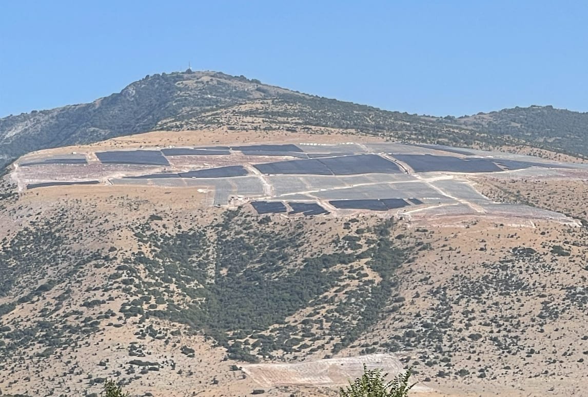 Hillside view of the Delfini solar project site in Greece, PV arrays laid out across graded plateaus below a mountain ridge