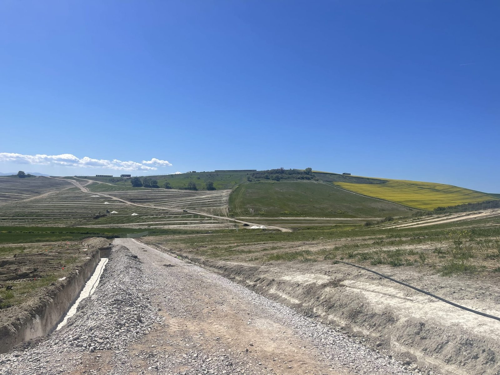 Gravel access road and an open cable trench under construction across a PV site in Greece, graded terraces on the hillside behind